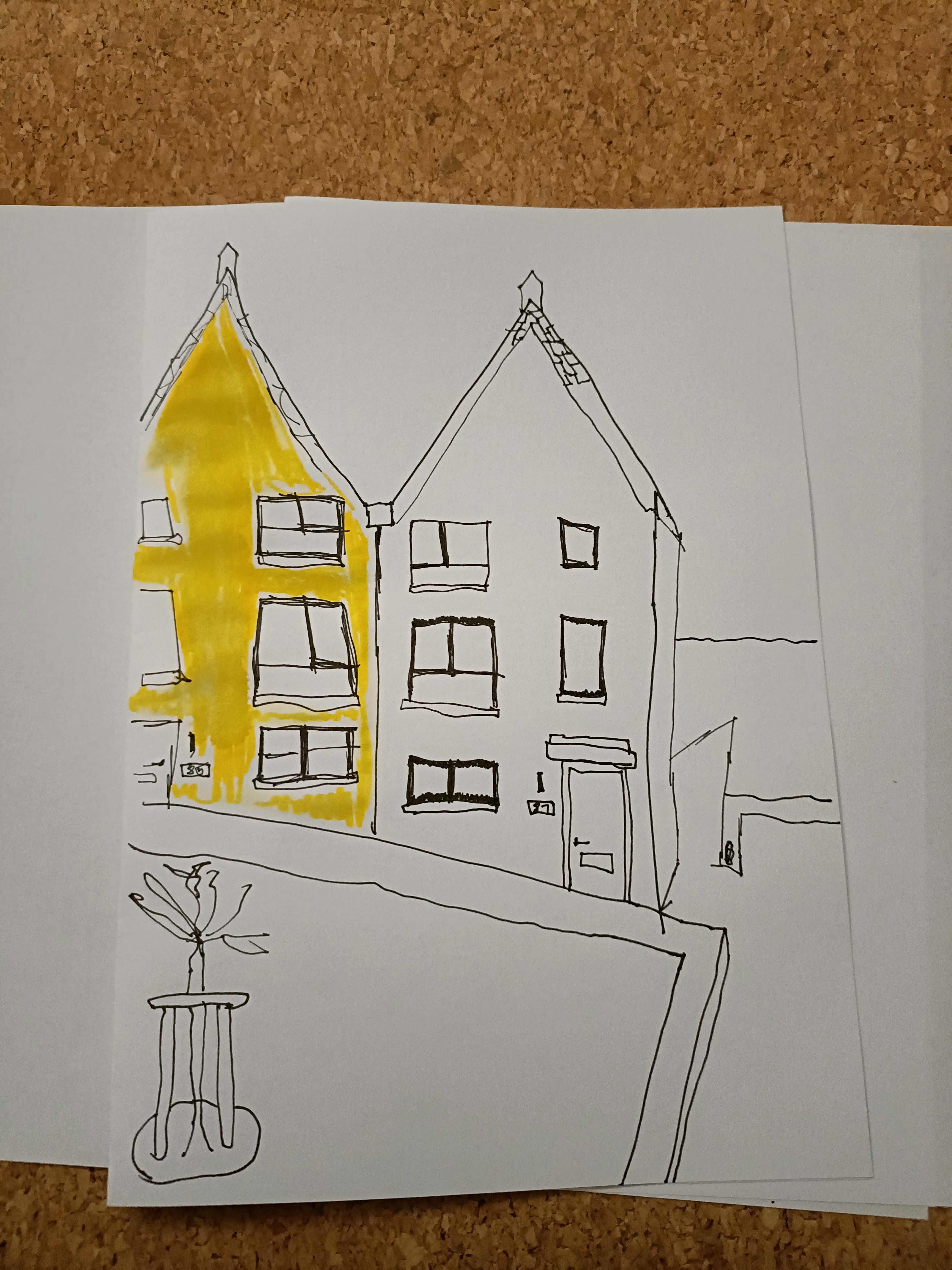 A simple ink drawing of two houses with a splash of yellow watercolor on the left one, sitting on a white sheet of paper. A sketch of a small sculpture is visible in the foreground.