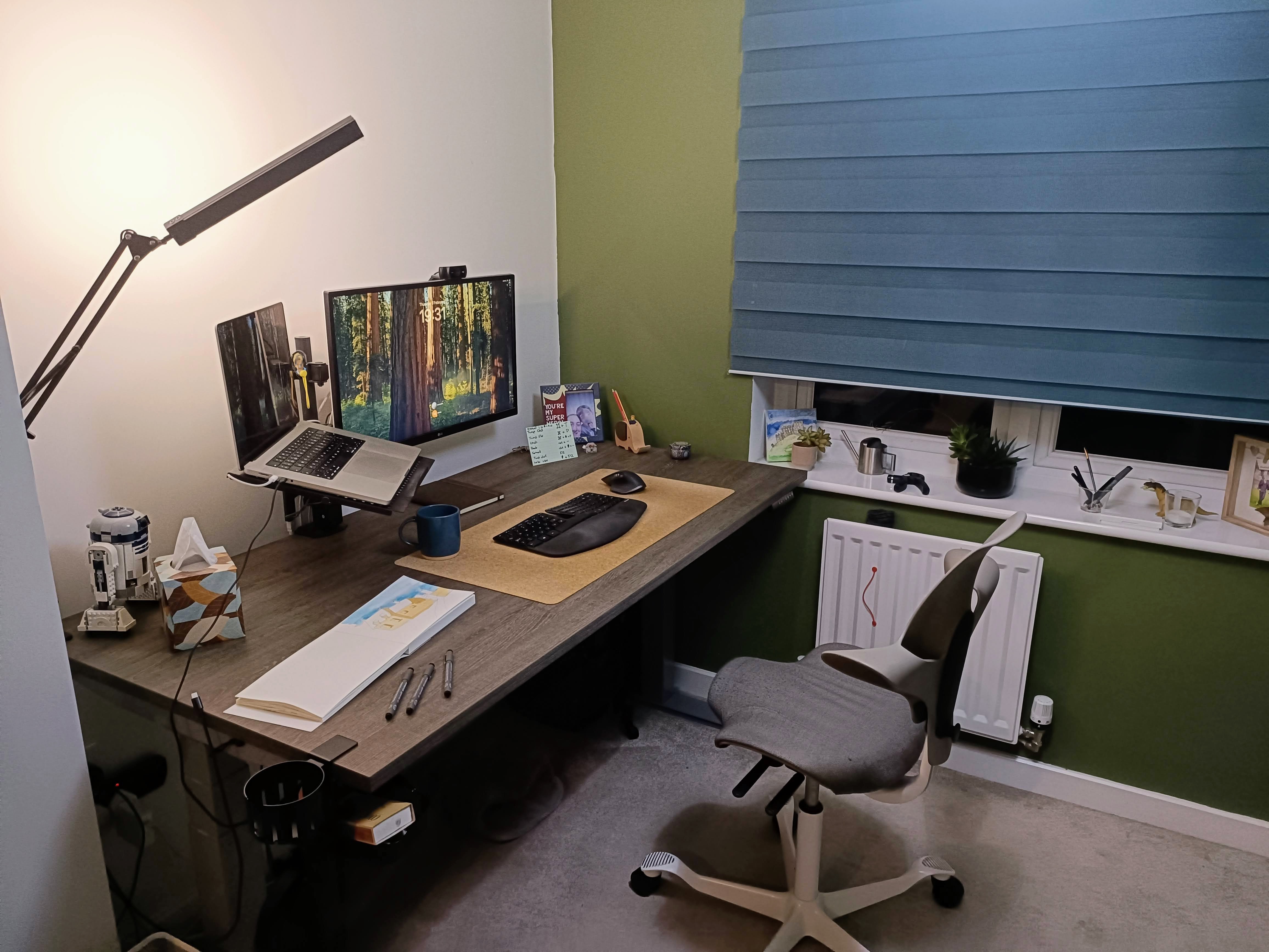 A modern home office setup featuring a desk with dual monitors, a laptop, a black keyboard, and a blue coffee mug. The desk is decorated with stationery, a book, and a tissue box. A comfortable office chair is positioned next to the desk, and there is a window with a blue blind and small plants on the sill.